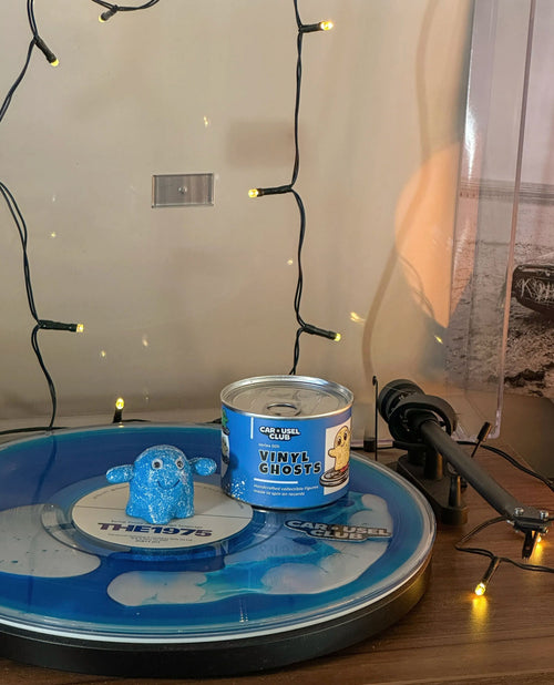 A blue vinyl record with a small blue ghost figurine and a can of Vinyl Ghosts, displayed on a turntable with warm string lights in the background.